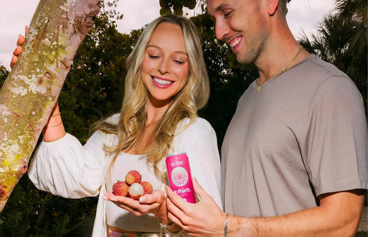 Couple outdoors holding fresh lychee fruit and a pink Kava by Mitra9 Seltzer can, natural tropical setting at sunset highlighting botanical-inspired calm drink.